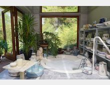 Helles Badezimmer mit einladender runder Badewanne, umgeben von Pflanzen, großen Fenstern mit Blick ins Grüne und Regalen mit dekorativem Keramikgeschirr.