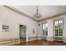 Ein eleganter Raum mit weiß gestrichenen Wänden, hohen Decken und verziertem Stuck. Über dem klassischen Holzboden hängt ein großer Kronleuchter, während große, rechteckige Fenster Tageslicht einlassen; eine offene Tür führt in einen angrenzenden prunkvoll dekorierten Raum.