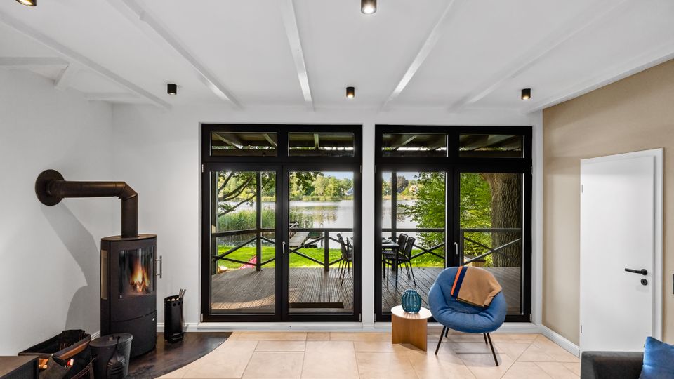 Heller Wohnraum mit weißer Balkendecke, Kaminofen an der linken Wand und großflächigen, schwarz gerahmten Terrassentüren. Durch die Glasfront ist eine Holzterrasse mit Sitzmöbeln und Blick auf einen See mit Bäumen und Ufergrün zu sehen.