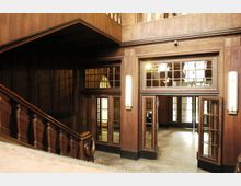 Holzvertäfelter Innenraum mit einer Holztreppe und mehreren verglasten Doppeltüren, die zu einem weiteren Raum führen. Oben sind Geländer und Holzdetails sichtbar, die den klassischen Stil der Architektur betonen.