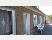 Außenansicht eines langgestreckten Gebäudes mit verputzter, beiger Fassade und mehreren großen Fenstern sowie Glastüren. Vor dem Gebäude stehen weiße Plastikstühle auf einer gepflasterten Terrasse.