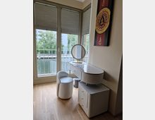 Ein heller Raum mit hellem Holzboden, einem Schminktisch mit ovalem Spiegel, passenden goldenen Details und einem weißen Polsterstuhl. Im Hintergrund sind bodentiefe Fenster mit Blick auf Bäume und ein Gewässer zu sehen, an der Wand hängt ein dekoratives Kunstwerk mit buddhistischen Motiven.