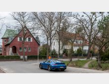 Straßenszene in einer Wohngegend mit einem roten Backsteinhaus mit grünem Dach und weißen Fensterläden, einem weißen Gebäude im Hintergrund und kahlen Bäumen. Im Vordergrund steht ein blauer Sportwagen geparkt an einer asphaltierten Straße mit angrenzendem Gehweg.