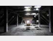 Verlassener Dachboden eines heruntergekommenen Gebäudes mit Betonpfosten, schrägen Holzbalken und mehreren kreisförmigen Öffnungen auf dem Boden. Graffiti bedecken die Wände, und Licht fällt durch Löcher in der Decke.