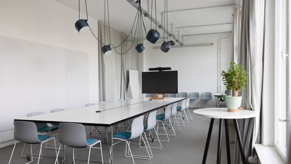 Heller, moderner Konferenzraum mit langem weißen Besprechungstisch und mehreren Stühlen auf Rollen, darüber hängende schwarze Pendelleuchten an sichtbaren Decken-Schienen. An der Stirnseite steht ein großer Bildschirm, rechts sorgen hohe Fenster mit Vorhängen und ein Pflanztopf auf einem runden Beistelltisch für Tageslicht und Akzente.