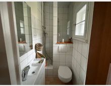 Kleines, schmal geschnittenes Badezimmer mit weißen Wandfliesen, Waschbecken mit Spiegel links und wandhängender Toilette rechts unter einem Fenster mit Milchglas. Am Ende des Raums befindet sich eine geflieste Dusche ohne Kabine, mit Handbrause und Ablageflächen.