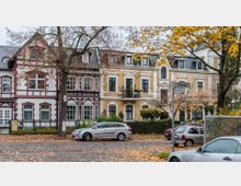 Straßenszene mit historischen Wohnhäusern, ergänzt durch Fachwerk- und Backsteinfassaden; im Vordergrund geparkte Autos auf einer gepflasterten Straße und buntes Herbstlaub von umliegenden Bäumen. Schild zeigt Straßennamen „Feldstraße“ und „Boninstraße“.