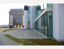 Moderne Bürogebäude mit großen Glasfassaden und abgerundeten weißen Säulen. Im Hintergrund ein weiteres Gebäude mit Ziegelfassade und großen Fenstern, umgeben von einem gepflasterten Gehweg, Grünflächen und geparkten Autos.