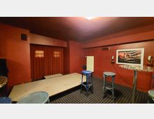 Kleiner Veranstaltungsraum mit roten Wänden und dunklem Teppichboden, einem erhöhten Bühnenbereich mit geschlossenem roten Vorhang, mehreren hohen Barhockern und Tischen sowie einem Heizkörper und einem gerahmten Kunstwerk an der Wand.