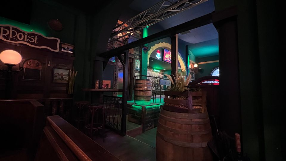 Innenraum einer dunklen Bar mit grüner Akzentbeleuchtung, Holzbänken und kleinen Tischen sowie mehreren großen Holzfässern im Vordergrund. Ein großer Rundbogen führt in einen blau beleuchteten Bereich mit Barwand, Bildern und Neonanzeigen.