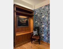 Holzgetäfelter Schrank mit integriertem Gemälde und beleuchtetem Fach, daneben ein dunkler Holzstuhl mit dekorativem Kissen. Der Raum hat einen Holzboden und eine auffällige Tapete mit blauem Hintergrund und stilisierten Tiermustern.