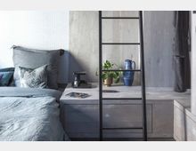 Modernes Schlafzimmer mit grauem Bett und minimalistischen Möbeln. Auf einem Nachttisch stehen eine Zimmerpflanze in einem goldenen Topf, eine blaue Keramikvase und eine Lampe; im Vordergrund ist eine schwarze Leiter zu sehen.
