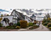 Wohnstraße mit Einfamilienhäusern und kleinen Vorgärten, links ein Wegweiser, im Hintergrund ein modernes Hochhaus. Bewölkter Himmel und herbstliche Vegetation umrahmen die Szene.
