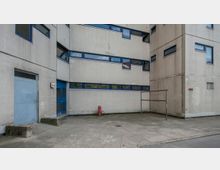 Eckansicht eines schlichten Gebäudes mit hellen, quadratischen Kacheln an der Fassade und blauen Fenstereinfassungen. Im Vordergrund befindet sich ein betonierter Hof mit einem roten Hydranten und einer Metallkonstruktion.