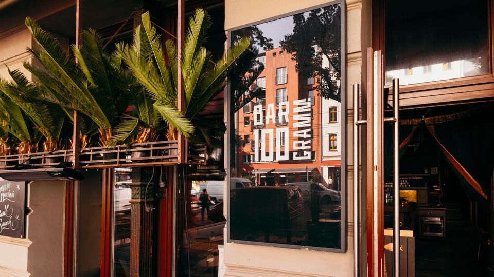 Eingangsbereich einer Bar mit großen Glasfenstern, Metalltürgriffen und einem großen Schaukastenplakat mit der Aufschrift „BAR 1000“. Links stehen hohe Pflanzen in einem erhöhten Pflanzkasten vor der Fassade; in der Glasscheibe spiegeln sich Straße und gegenüberliegende Gebäude.
