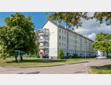 Mehrstöckiges Wohngebäude mit hellbeiger Fassade und metallenen Balkonen, umgeben von Grünflächen, Bäumen und Gehwegen. Im Vordergrund ist eine Straßenecke mit Straßenschildern und geparkten Autos zu sehen.