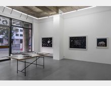 Innenraum einer modernen Kunstgalerie mit weißen Wänden und Beton-Decke. An den Wänden hängen gerahmte Bilder mit abstrakten Motiven und Texten, und ein schlichter Tisch mit schwarzen Metallbeinen steht im Vordergrund, darauf liegen geöffnete Kunstbücher. Durch die großen Fenster ist eine Straße mit Gebäuden sichtbar.