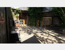 Holzterrasse im Freien mit einer kleinen Holzbühne im Hintergrund, dekoriert mit einem Pult, auf dem ein Symbol mit den Buchstaben „SC“ zu sehen ist. Links und rechts stehen Bambuspflanzen und Holzgebäude, und das Gelände ist teils von Schatten bedeckt.