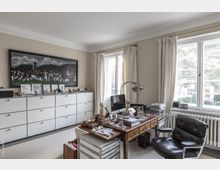 Helles Heimbüro mit cremefarbenen Wänden, zwei großen Fenstern mit weißen Vorhängen und einer Heizung darunter. Ein Schreibtisch aus Holz mit Büromaterialien und einem Computer steht vor einem schwarzen Bürostuhl, während an der gegenüberliegenden Wand eine weiße Kommode mit mehreren Schubladen und Dekorationsobjekten steht, über der ein Gemälde hängt.