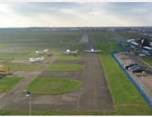 Kleiner Flugplatz mit abgestellten einmotorigen Flugzeugen auf asphaltierten Flächen, umgeben von Rasen. Am rechten Bildrand befinden sich ein Parkplatz mit Autos und eine Reihe von wartenden Reisebussen, getrennt durch einen blauen Zaun.