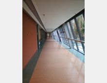 Langer Verbindungsgang mit modernen Glaswänden auf der rechten Seite, die einen Ausblick ins Freie bieten, und einer orangefarbenen Wand auf der linken Seite; der Boden ist aus beigem und grünem Fliesenmaterial.