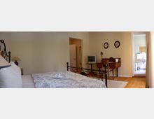 Schlafzimmer mit einem schmiedeeisernen Bett, hellem Bettwäsche-Design, einem kleinen Fernseher auf einem antiken Schreibtisch und Holzboden. Im Hintergrund befindet sich ein offener Zugang zu einem Badezimmer mit weißen Wandfliesen und einem modernen Waschbecken.