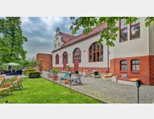 Außenansicht eines historischen Gebäudes mit roten Backstein- und Putzfassaden sowie großen Rundbogenfenstern. Im Vordergrund befindet sich ein Gartenbereich mit Liegestühlen, Sitzgelegenheiten und Sonnenschirmen, umgeben von Rasenflächen und Bäumen.