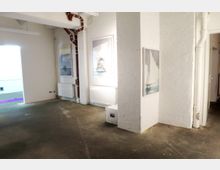 Ein heller, minimalistischer Raum mit weißen Wänden und Sichtmauerwerk, dekoriert mit großformatigen Kunstwerken. Sichtbare Heizungsrohre verlaufen entlang der Wände, und der Boden besteht aus grauem Beton.