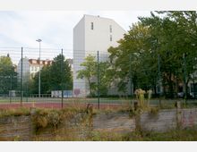 Ein Sportplatz mit rotem Belag, umgeben von einem hohen Metallzaun, im Vordergrund eine verwilderte Böschung mit Holzbegrenzung. Im Hintergrund steht ein weißes, modernes Gebäude neben Bäumen und weiteren Wohnhäusern.