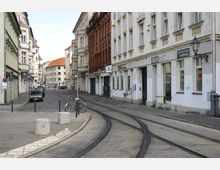Straßenszene mit Kopfsteinpflaster und Straßenbahnschienen, flankiert von mehrstöckigen historischen Wohn- und Geschäftsgebäuden. Im Vordergrund stehen Poller und ein Fahrrad, und geparkte Autos säumen die Straße.