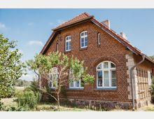 Backsteingebäude mit Satteldach und weißen Rundbogenfenstern, umgeben von vereinzelten Bäumen und Sträuchern in einer ländlichen Umgebung. Im Vordergrund befinden sich ein kleiner Baum und eine Wiese mit Pflanzen.