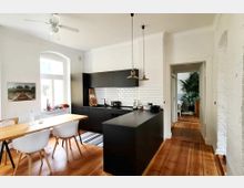 Moderne Küche mit Holzboden, weißen Wänden und einer schwarzen Küchenzeile mit weißen Fliesen im Metro-Stil. Ein Holztisch mit weißen Stühlen steht vor einem großen Fenster, im Hintergrund ist ein angrenzender Raum mit Pflanzen sichtbar.