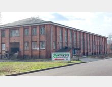 Backsteingebäude mit rechteckigen Fenstern, einem schrägen Dach und einer Rampe am Eingang; im Vordergrund befindet sich ein Schild mit der Aufschrift „Hyflexar Hydraulik-Technik“. Das Umfeld zeigt eine asphaltierte Straße und Grünflächen.