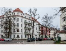 Ecke einer städtischen Wohnstraße mit mehrstöckigen Altbauten in hellen Farben, darunter ein weißes Gebäude mit roten Dachziegeln und grünen Balkonen. Im Vordergrund befinden sich geparkte Autos, Straßenlaternen und kahle Bäume.