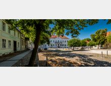 Gepflasterter Stadtplatz mit großen Laubbäumen und Baumschatten, umgeben von niedrigen historischen Gebäuden. Im Hintergrund steht ein weißes Rathaus mit rotem Satteldach und symmetrischer Fassade, links ein Gebäude mit der Aufschrift „Stadtbibliothek“.