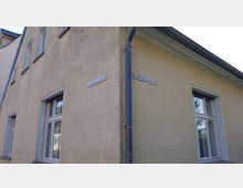 Ecke eines beigen Wohnhauses mit Schildern, die Straßennamen anzeigen, und mehreren Fenstern. Ein Regenfallrohr verläuft an der Ecke des Gebäudes entlang, und der Himmel ist im Hintergrund leicht bewölkt.