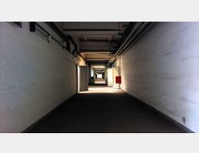 Langer, schmaler Flur mit weiß gestrichenen Wänden und sichtbaren Leitungen an der Decke. Im Hintergrund ist eine geöffnete Tür mit einem Schild „Museum“ zu sehen; an der rechten Wand hängt ein roter Kasten.