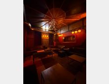 Gedimmter Lounge- oder Barbereich mit dunklen Lederbänken und Sesseln um niedrige Tische, warm beleuchtet von runden Wandlampen und einem zentralen Kronleuchter. Industrielle Details wie sichtbare Lüftungsrohre und Holzvertäfelung prägen den Raum, ergänzt durch eine rote Akzentwand mit großem Spiegel.