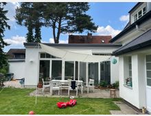 Einfamilienhaus mit weißer Fassade und modernen Fenstern; im Gartenbereich stehen weiße Gartenmöbel auf einer Terrasse unter einem Sonnensegel. Im Vordergrund Rasenfläche mit Spielzeug, im Hintergrund Bäume und benachbarte Gebäude.