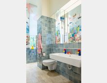 Modernes Badezimmer mit hellblauen Wandfliesen und hellem Fliesenboden. Die Wände sind mit meeresinspirierten Wandbildern dekoriert, es gibt ein großes Wandspiegelpaneel mit integrierter Beleuchtung, eine schwebende Toilette und ein weißes Waschbecken mit buntem Kinderspielzeug darauf.