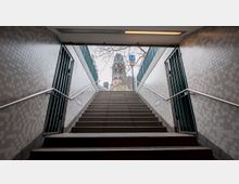 Treppenaufgang einer U-Bahn-Station mit gefliesten Wänden und Metallgeländern, der nach draußen führt; im Hintergrund ist die Turmruine der Berliner Kaiser-Wilhelm-Gedächtniskirche zu sehen. Ein blaues „U“-Schild markiert den Eingang zur U-Bahn.