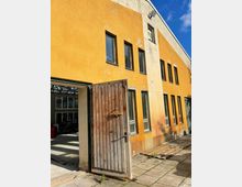 Gelbes, zweistöckiges Gebäude mit leicht verwitterter Fassade, mehreren rechteckigen Fenstern und einer offenen Holztür, die in einen Innenraum mit sichtbaren Geräten führt. Vorgelagert ist ein gepflasterter Boden, beleuchtet von Sonnenlicht.