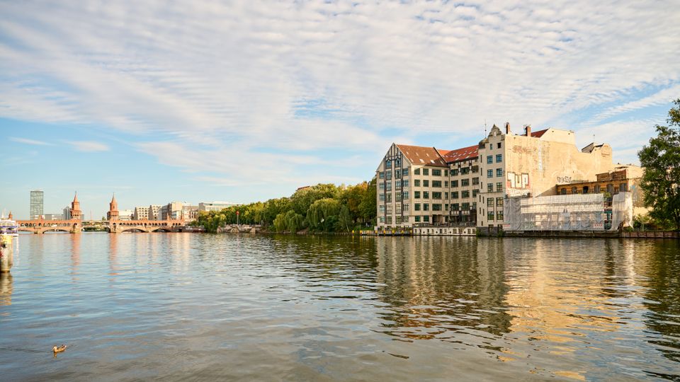 Blick über einen breiten Fluss auf ein Ufer mit einem großen, mehrstöckigen Gebäudeensemble mit Satteldächern und vielen Fenstern, teils mit Gerüst und Verkleidung. Im Hintergrund ist eine markante Brücke mit Türmen sowie weitere Stadtbebauung und eine Baumreihe entlang des Ufers zu sehen.