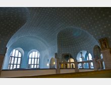 Ein Innenraum mit einer gewölbten, himmelblauen Decke, die mit kleinen floralen Mustern dekoriert ist. Große, halbkreisförmige Fenster und Rundbögen dominieren die Architektur, die durch Säulen und helles Steinmaterial ergänzt wird.