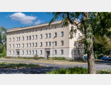 Wohngebäude im schlichten Baustil mit vier Stockwerken, hellbeigefarbener Fassade und regelmäßigen rechteckigen Fenstern. Im Vordergrund ein kleiner Grünstreifen mit Gehweg, Bänken und einem Baum, umgeben von parkenden Autos und einer Straße.