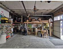 Werkstatt mit Regalen, Werkbänken und verschiedenen Werkzeugen. Holzplanken und Materialien sind auf einem erhöhten Lagerbereich gestapelt, der Raum ist von einer großen Garagentoröffnung mit Tageslichteinfall geprägt.