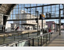 Heller, moderner Bahnhof am Alexanderplatz in Berlin mit großen Glasfassaden, überdachten Bahnsteigen und wartenden Personen. Im Hintergrund sind Gebäude des Stadtzentrums sichtbar.