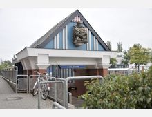 Außenansicht des Eingangs zum U-Bahnhof Zitadelle mit spitzem Dach, Zierelementen in Blau und Weiß und einer Statue auf der Fassade. Im Vordergrund sind Fahrradständer, ein Gehweg mit Geländer und grüne Büsche zu sehen.