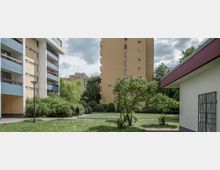 Wohnsiedlung mit mehreren mehrstöckigen Gebäuden, darunter ein gelb-orangefarbenes Hochhaus und ein Wohnhaus mit blauen Balkonfassaden. Im Vordergrund eine Grünfläche mit Bäumen, Sträuchern und einem Gehweg, auf dem zwei Personen spazieren.