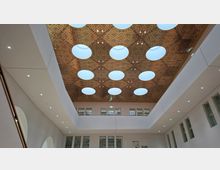 Innenansicht eines modernen Atriums mit hoher Decke aus perforierten Holzpaneelen und mehreren runden Oberlichtern. Umgeben von hellen Wänden mit integrierten Spotlights sowie Fensterreihen und Galerien auf mehreren Ebenen.